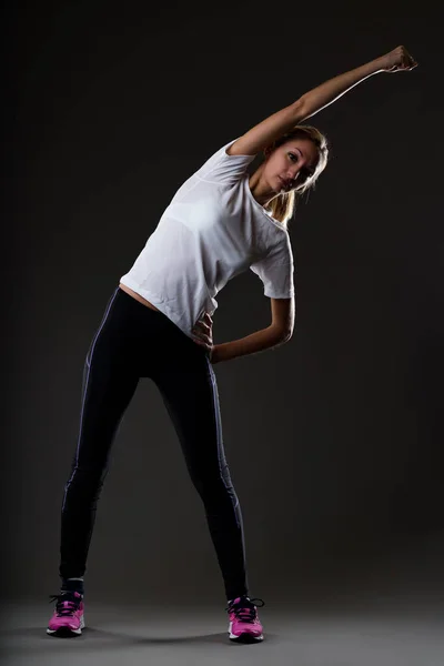 ブロンドの女性は 黒の背景に 黒のフィッティングパンツ フクシアスニーカー 白いTシャツを着て運動 手を腰に横に曲げ腕を伸ばす — ストック写真