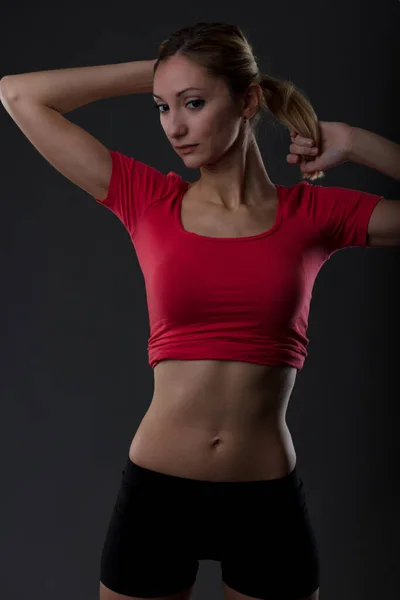 健康的女人用体操锻炼来保持健美的身体 体育活动反映了决心和责任 穿着红衬衫 扁平腹部 黑色背景短裤的金发姑娘的画像 — 图库照片