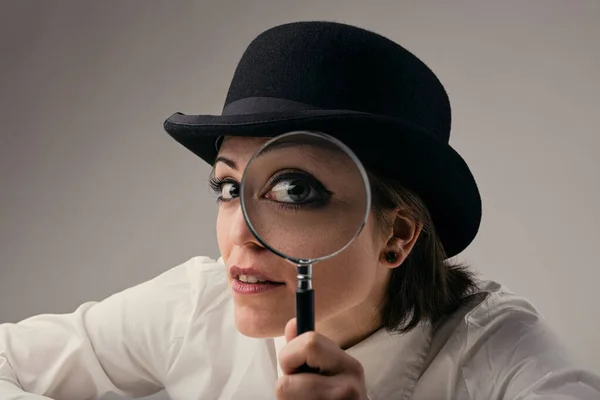 Intense close-up of a woman\'s eye, peering through a magnifying glass while sporting a bowler hat. Private investigator vibes, potential data breach, invasion of privacy. It\'s anxiety-inducing