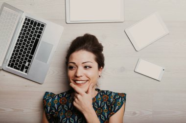 A woman smiles, she's had a great idea! This will solve the problem, please the customer, and profit her. Surrounded by laptop, tablet, and various smartphones with blank screens, in a direct overhead clipart