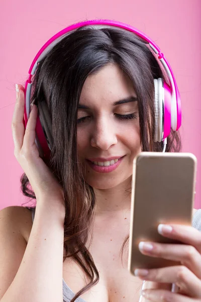 年轻的女人闭上眼睛 微笑着 在粉色的背景下听着粉色的耳机 沉浸在音乐或播客中 — 图库照片