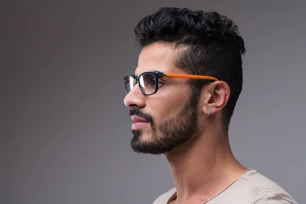 Attractive Middle Eastern man, side-bust shot. Wearing a beige shirt, colorful glasses, an earring. His fitness, black hair, and groomed beard are striking
