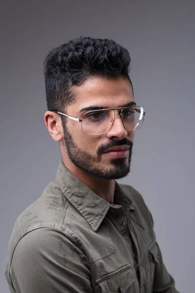 Three-quarter bust portrait of a fit, attractive Middle Eastern man. His black hair and neat beard compliment his green shirt and metallic glasses