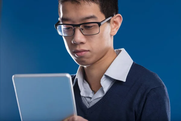 一个全神贯注的亚洲人 一边看着平板电脑上的新闻 一边戴眼镜 在蓝色的背景下 他与屏幕 电子邮件或摄像头视图 进行了接触 — 图库照片
