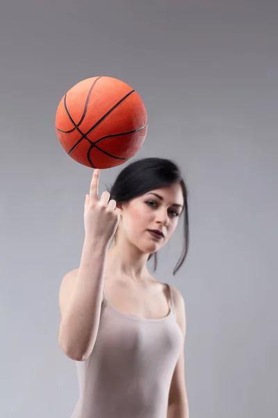 有吸引力的运动的年轻女子平衡一个篮球在她的指尖 而看着相机与一个认真周到的表达在灰色演播室背景 — 图库照片