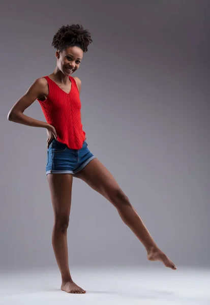 playful young woman with brown skin, afro hair, red shirt, and jean shorts that bare her beautiful legs. Barefoot, pointing toe like a dancer or footballer. Smiling sweetly, she\'s happy and feels good