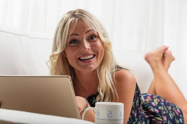 在她的沙发上 一个快乐的金发碧眼的女人带着平板电脑开心地微笑着 她可能是在购物 沉迷于电视节目或观看社交媒体视频 — 图库照片