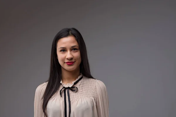 Half-length portrait of a proud, intelligent, confident young Filipina woman. She wears a beige blouse adorned with a black ribbon. Grey background