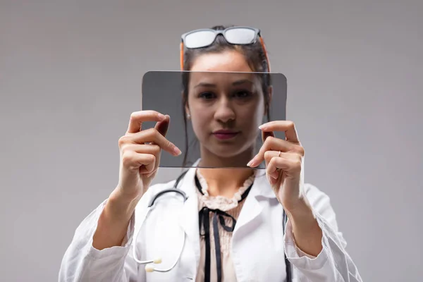 Female doctor analyses YOUR COPYSPACE data on a digital tablet. The futuristic transparent screen is empty for your input. Glasses worn as a headband, she is serious