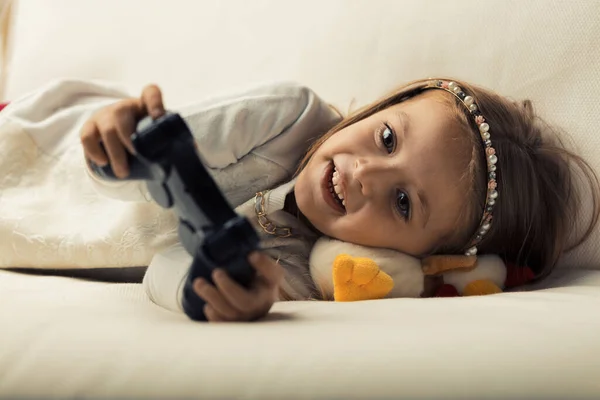 A little girl enjoys video gaming on her home sofa with a controller, under her parents' watchful eyes