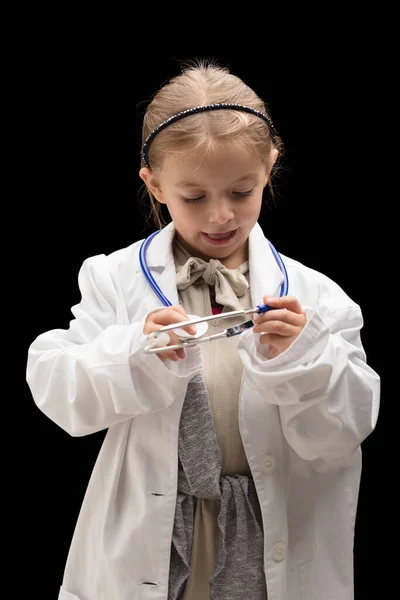 小さな女の子は 黒い背景に対してオーバーサイズの白いコートとスタチオスコープの医者であるふりをする かわいいブロンド 彼女は将来獣医になることを目指しているかもしれません — ストック写真