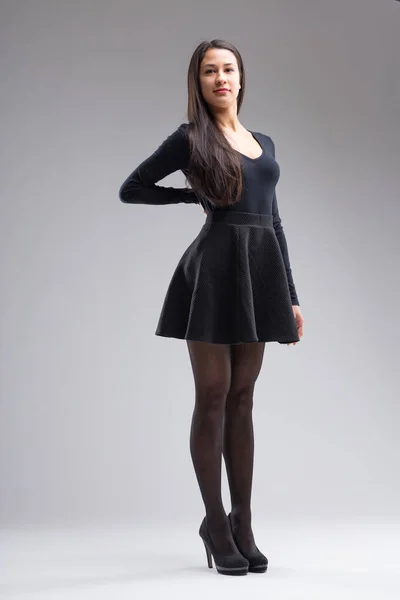 A full-length portrait of a young, tall, and beautiful Filipino woman. Dressed in black from long sleeve top, miniskirt, to sheer stockings. Posing proudly on a neutral gradient background