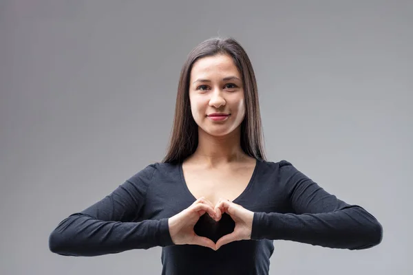 Half-bust frontal portrait of a young woman forming a heart with her fingers. \'I love you! I love all this! I adore this situation or thing.\' She wears a black dress and has long straight dark hair, s