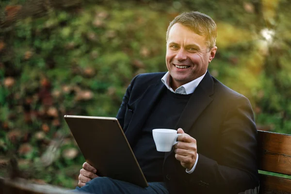 中年男性はコーヒーを飲みながらオフィスガーデンでタブレットを使用し データ分析と調整を可能にします 公園でニュースを読む退職者に最適です — ストック写真