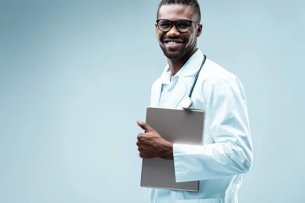 Smiling Medical doctor, black with neat beard, uses digital tools for quality patient care. Wields a tablet, white coat, stethoscope, highly educated, handsome