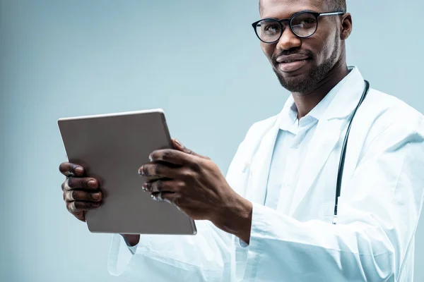 Modern male doctor, black, neat beard, using digital tech. Holds a tablet, white coat, stethoscope, multiple degrees and handsome