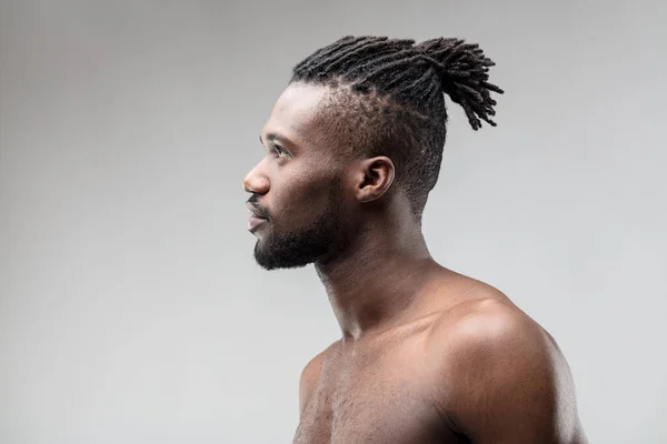 Side view, head and shoulders of a black man, chest and back bare, and a neat beard. Confident in his attractiveness, he poses on a gray background