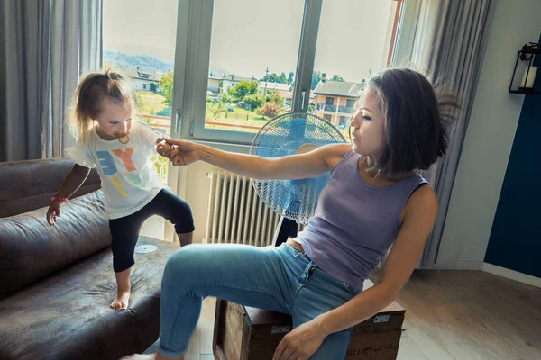 Mom and small daughter dance and fly with a fan in the living room, forgetting the heat and seeking refreshment joyfully