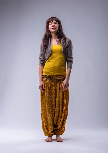 Full-length portrait of an adult woman against a neutral backdrop, dressed in warm yellow and beige, with puffed trousers and barefoot. Her face urges: stop wasting time