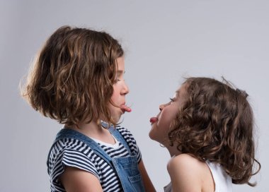 Two young girls, aged 5-7, teasingly stick out their tongues. Likely friends or sisters, they face each other making faces, dressed casually. They laugh joyfully clipart