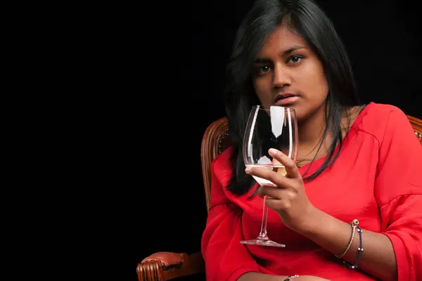 Celebrating the art of wine, an Indian woman swirls quality white wine, seated on a timeless leather and wood chair. Dressed in a vibrant red, she\'s set against a dramatic black background