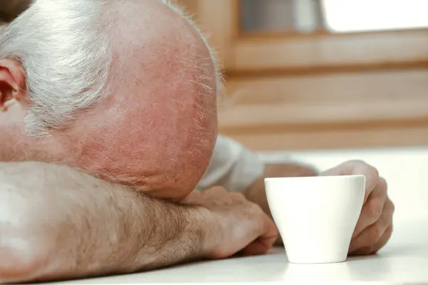 stock image With dawn breaking, an old man grapples with fatigue, sipping his coffee. His daytime job is at risk due to sleeplessness. Earnings don't match the rising retirement age. Medical assistance is essenti