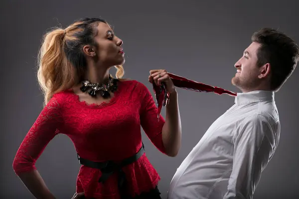 Woman in red with playful smirk holds a man\'s tie, suggesting control