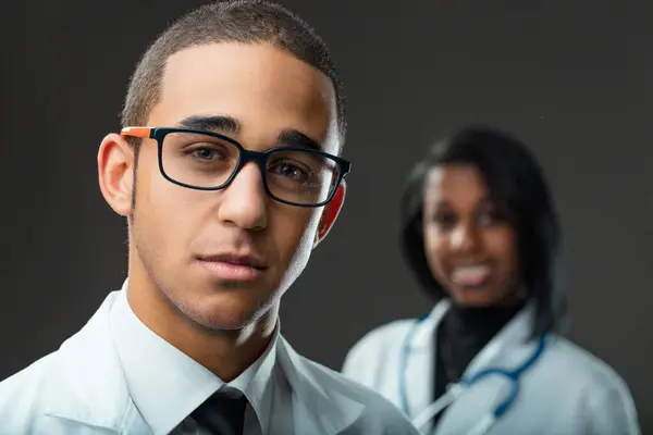眼鏡の若い男性医学生は 女性医師が背景に微笑んでいる間 焦点を当てています — ストック写真