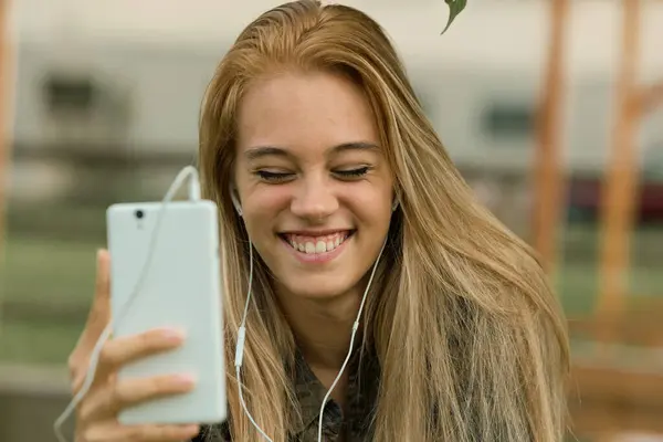 彼女はビデオ通話を楽しんでいるので 若い女性に放射性微笑み イヤホンでスマートフォンを保持 ストックフォト