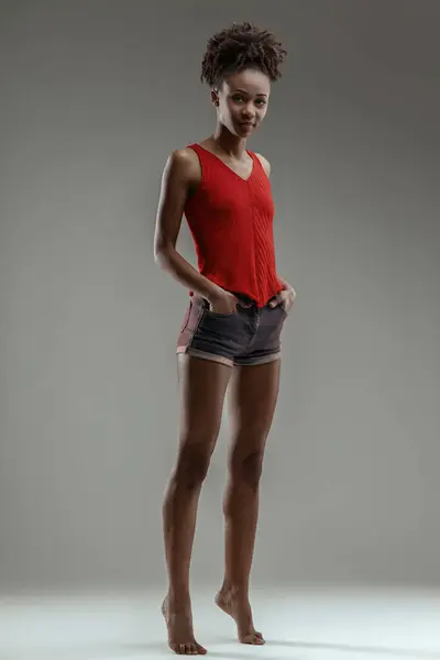 若い美しい黒人女性は 赤い袖口とデニムショートパンツでハンズオンシップ裸足を立っています ストック写真