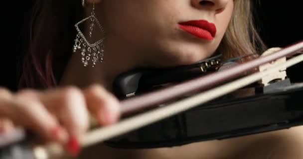 视频中 一个从小提琴家的脸上到躯干的垂直平底锅显示了她在演奏一把黑色电小提琴 她穿着黑色无袖连衣裙装饰着红色口红 耳环和金发 背景是黑色的 — 图库视频影像