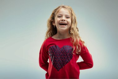 Young girl with curly blonde hair laughs joyously, wearing a red sweater adorned with a large heart pattern, her happiness contagious clipart