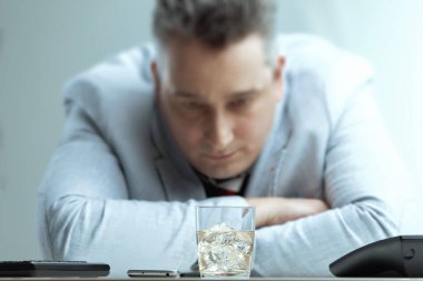 middle-aged man in a light suit stares intently at a glass of whiskey on his desk, battling the urge to drink. His somber expression reflects the internal struggle and the potential consequences clipart