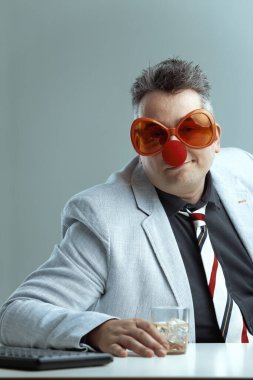 middle-aged man in a light suit and striped tie wears oversized orange glasses and a red clown nose, holding a whiskey glass. His playful, ridiculous appearance underscores the folly of drinking at wo clipart