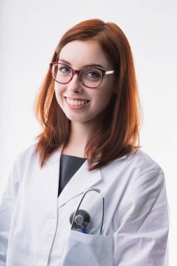 Smiling young woman with auburn hair wears glasses and a white lab coat, with a stethoscope in her pocket. Her friendly demeanor and professional appearance convey competence and approachability clipart