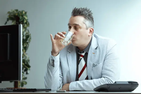 Stock image Man with spiky gray hair, light gray blazer, dark gray shirt, and striped tie, holds a glass of alcohol with a smug look, believing it normal to drink at work, office setting