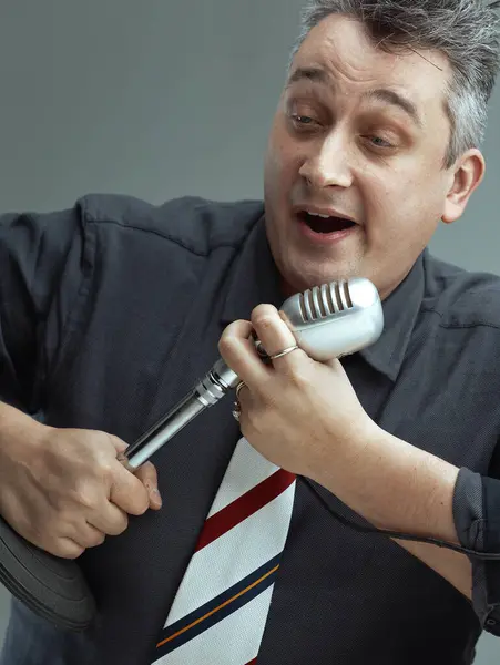 stock image man, dressed in a dark shirt and striped tie, sings energetically into a vintage microphone. His expressive demeanor and enthusiastic posture show his deep involvement in the song
