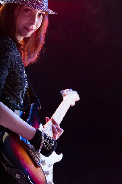 stock image Smiling auburn-haired woman wearing a glittery hat and dark top plays an electric guitar. Her confident stance and joyful expression on stage emphasize her love for music