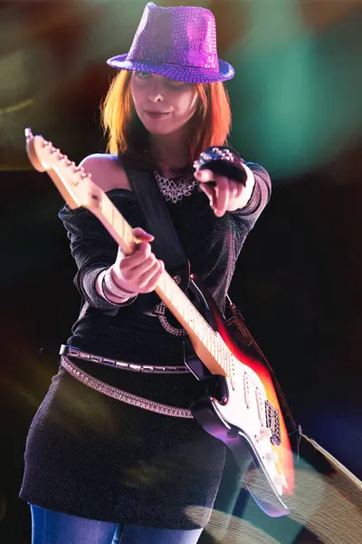 stock image  joyful young woman with red hair, in a glittering black ensemble and sequined hat, skillfully plays her electric guitar, stage lights casting colorful reflections around her