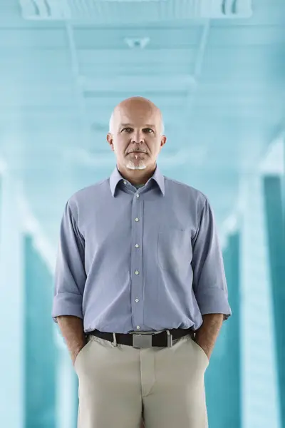 stock image Bald man with white goatee and light blue shirt stands with hands in pockets, looking confidently at the camera. The blue corridor behind him highlights his calm and professional demeanor