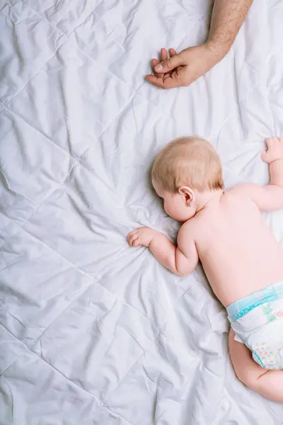 stock image Baby sleeps peacefully on a white quilt, his tiny hand grasping the fabric. Nearby, a man's hand, likely the father, rests exhaustedly. The scene reflects the shared fatigue and tender bond