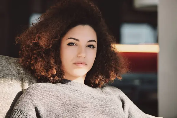 stock image beautiful woman with curly hair sits assertively in her living room, exuding confidence and control over her life. Her calm, composed expression reflects her inner strength and self-assuredness