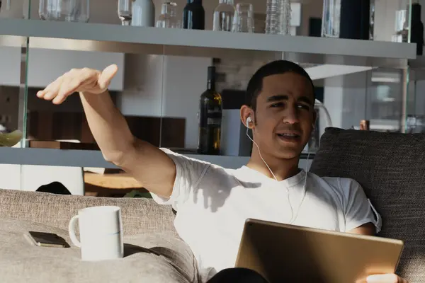 stock image oung man in a white t-shirt, wearing earphones, is excited about the music he's listening to. Seated on a sofa, holding a tablet, he gestures energetically. A mug and phone rest beside him