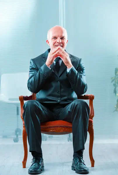 stock image bald man in a dark suit sits confidently, hands clasped together, exclaiming 'excellent.' He represents a ruthless multinational owner focused solely on shareholder value, disregarding employee welfar