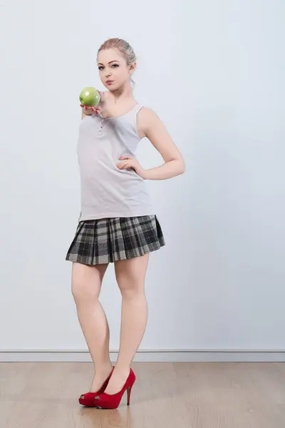 stock image young woman, dressed in a plaid skirt and red heels, holds a green apple with a knowing expression. The apples metaphorical meaning suggests a playful nod to temptation and decision 