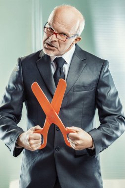 Man in suit displays a mock sadistic pleasure in holding large scissors, symbolizing severe corporate downsizing practices clipart