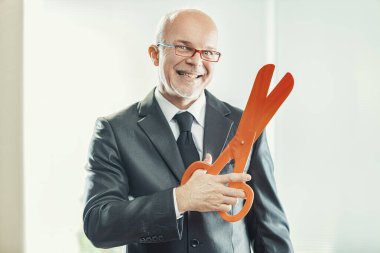 Smiling businessman holds large scissors, mockingly representing the cost-cutting measures often disguised as valuing personnel clipart