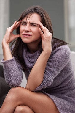 Young woman sits at home, touching her head in pain, looking tired and sick. She seems unhappy and worried, reflecting common health struggles clipart