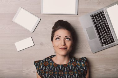 Young woman lies on a wooden floor surrounded by digital devices, lost in thought and gazing up, capturing the essence of the modern digital age clipart