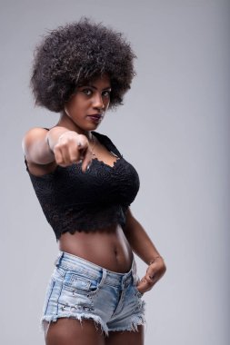 Young woman with afro hair pointing her finger at the viewer, displaying a serious expression. Dressed in jeans shorts and a black top, exuding confidence and assertiveness clipart